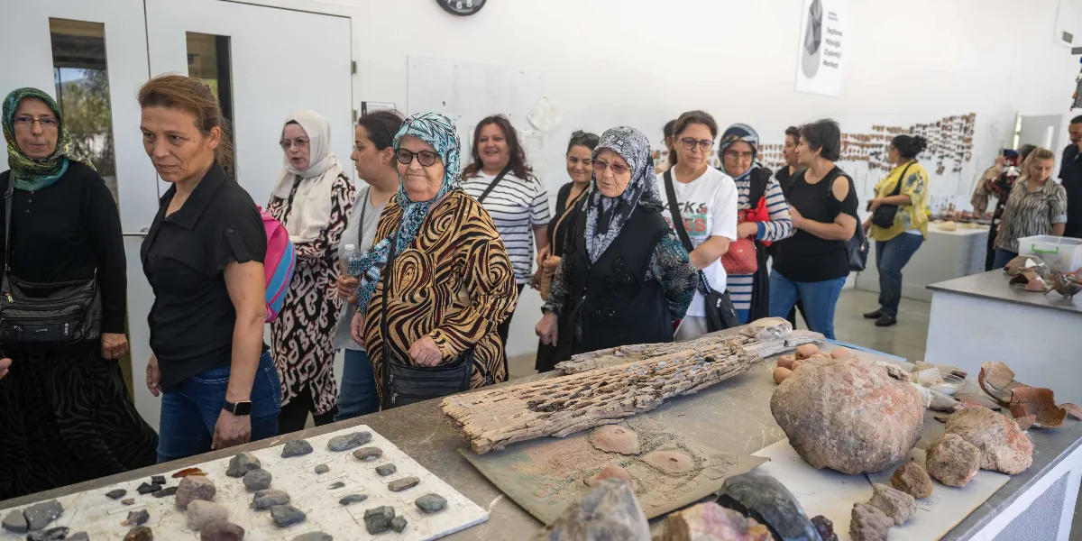 Aydınlık bir sergi salonunda, masanın üzerindeki arkeolojik buluntuları inceleyen bir grup kadın ziyaretçi. Masanın üzerinde çeşitli taş parçaları, eski bir ahşap kalıntı ve büyük bir toprak çömlek parçası sergileniyor. Ziyaretçiler, masanın etrafında toplanmış, dikkatle eserleri inceliyor.