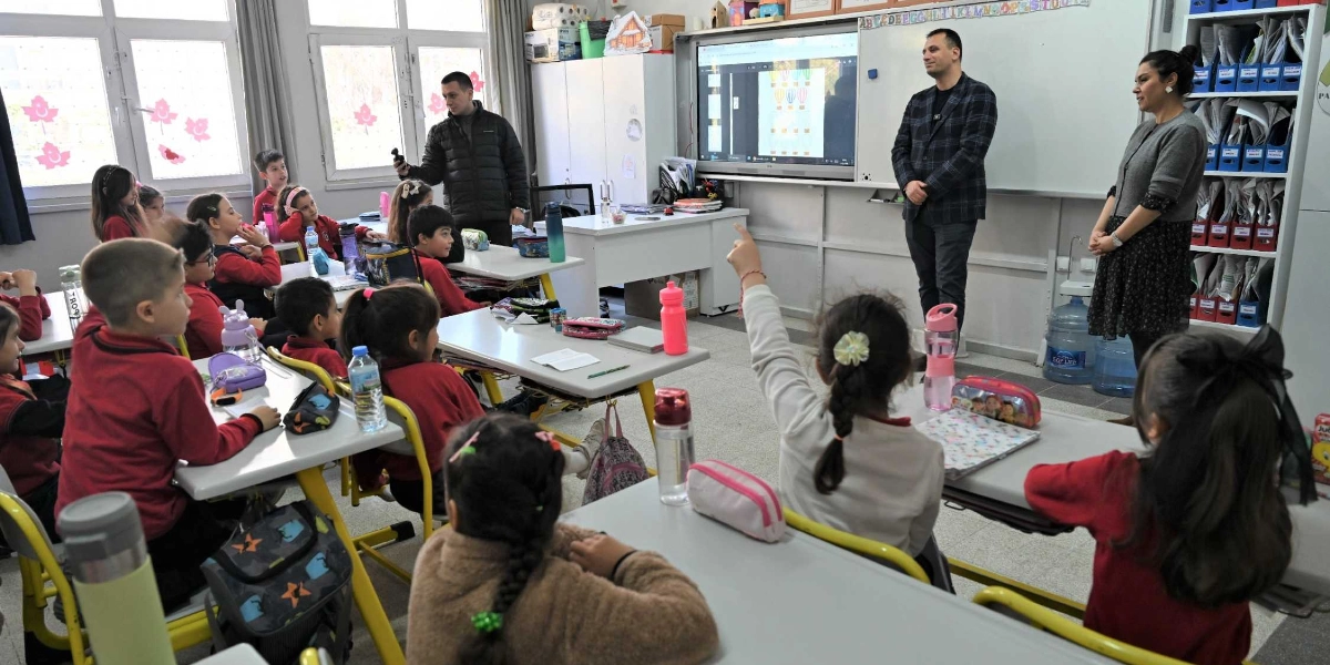 Sınıf ortamında, kırmızı üniformalı ilkokul öğrencileri sıralarında otururken, ön tarafta duran bir erkek ve bir kadın yetişkin onlara hitap ediyor. Sınıfın önündeki akıllı tahtada balon görselleri açıkken, bir öğrenci parmağını kaldırarak söz istiyor. Sınıfın sol tarafında ayakta duran başka bir erkek ise elinde bir cihazla ortamı görüntülüyor.