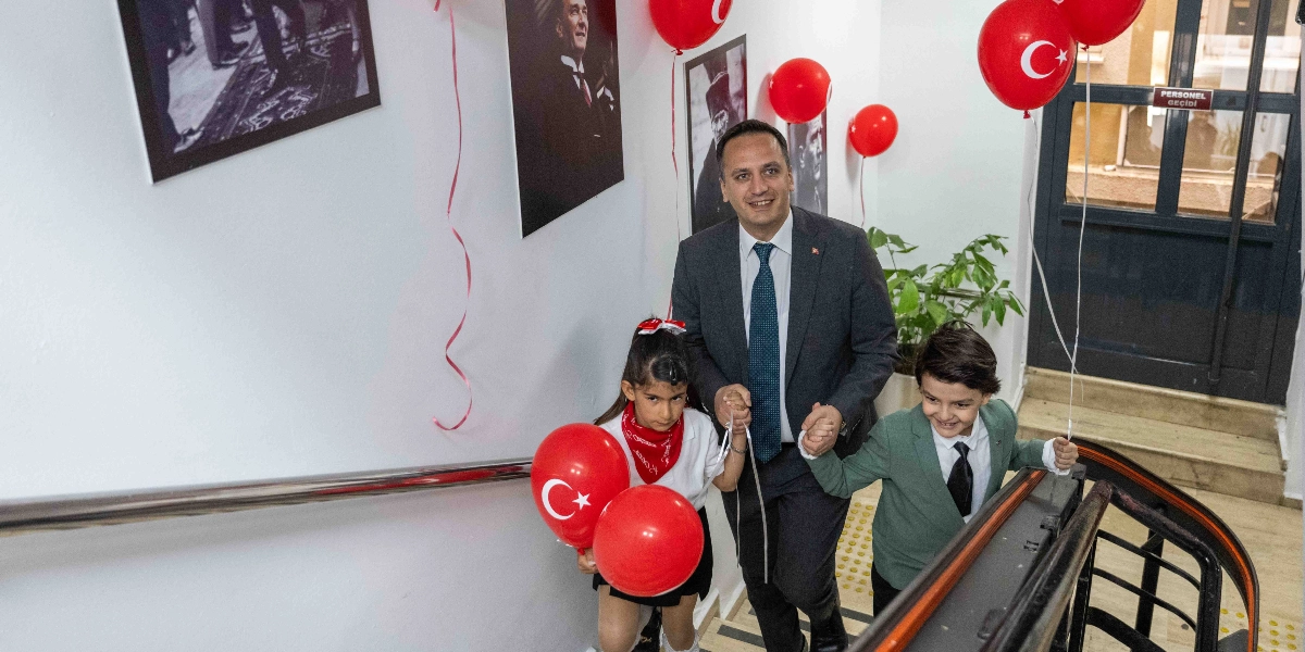 Bornova Belediye Başkanı Mustafa İduğ, 23 Nisan Ulusal Egemenlik ve Çocuk Bayramı kapsamında makamına eşlik eden bir kız ve bir erkek çocukla birlikte merdivenlerden gülümseyerek iniyor. Başkan koyu renk takım elbise ve kravat giymiş, çocukların ellerini tutuyor. Kız çocuğu beyaz bir bluz ve kırmızı fular takmış, elinde iki adet Türk bayraklı kırmızı balon tutuyor. Erkek çocuk ise yeşil bir ceket ve kravat giymiş, elinde bir adet Türk bayraklı kırmızı balon tutuyor. Duvarlarda Atatürk fotoğrafları asılı ve ortam kırmızı balonlarla süslenmiş durumda.