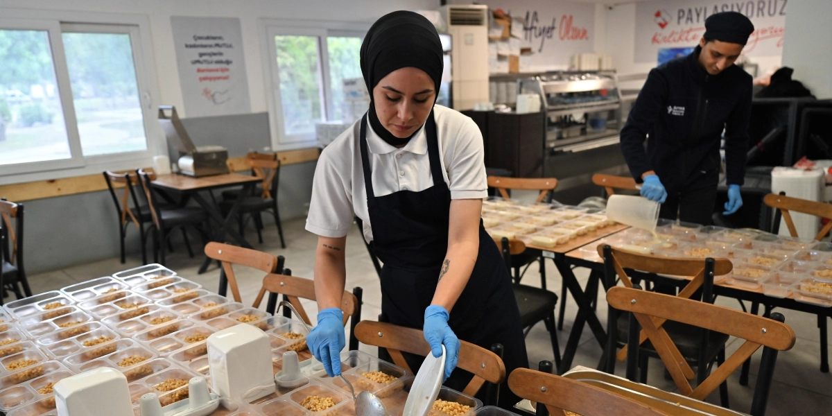 Beyaz önlük ve siyah önlük giymiş, başında siyah bir başörtüsü bulunan genç bir kadın, endüstriyel bir mutfakta plastik kaplara yemek paylaştırıyor. Kadının ellerinde mavi koruyucu eldivenler bulunuyor ve önündeki masada çok sayıda yemek dolu kap sıralanıyor. Arka planda, benzer kıyafetler giymiş bir başka çalışan daha yemek paketleme işlemine devam ediyor. Mutfağın duvarında 