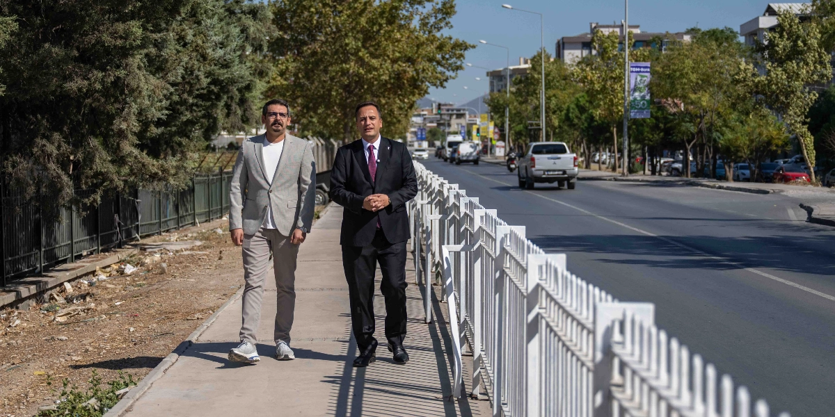 Güneşli bir günde, şehir içi bir caddenin kenarındaki kaldırımda yan yana yürüyen iki erkek. Sağ tarafta, koyu renk takım elbise ve bordo kravat giymiş, ellerini önünde birleştirmiş bir adam; sol tarafta ise gri ceket, beyaz tişört ve bej pantolon giymiş, bıyıklı başka bir adam yer alıyor. İkilinin sağ tarafında beyaz metal bir korkuluk, sol tarafında ise ağaçlıklı bir alan bulunuyor. Arka planda ise trafik akışının olduğu asfalt bir yol ve ağaçlar görülüyor.