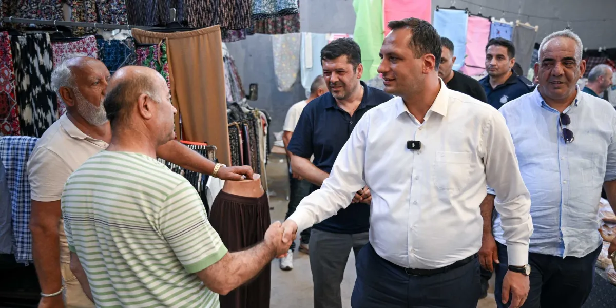 Bornova Belediye Başkanı Ömer Eşki, bir pazaryerini ziyaret ederek esnafla tokalaşıyor. Beyaz gömlekli ve lacivert pantolonlu olan belediye başkanı, tezgahların önünde duran esnafla sohbet ederken, çevresinde diğer belediye yetkilileri ve güvenlik görevlileri yer alıyor. Arka planda ise askıda kıyafetlerin sergilendiği bir pazar tezgahı görülüyor.