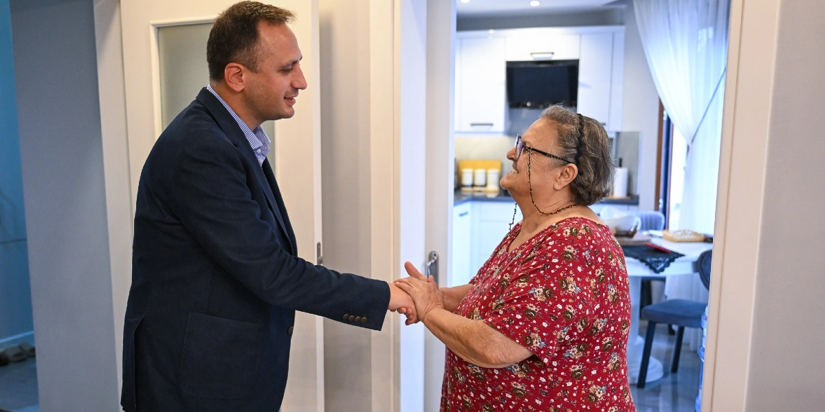 Koyu renk ceket giymiş bir erkek, ev ortamında karşılaştığı yaşlı bir kadının elini tutarak onunla samimi bir şekilde selamlaşıyor. Kadın, üzerinde çiçek desenli kırmızı bir bluzla gülümseyerek erkeğe bakıyor. Arka planda ise modern bir mutfak ve yemek alanı görünüyor.