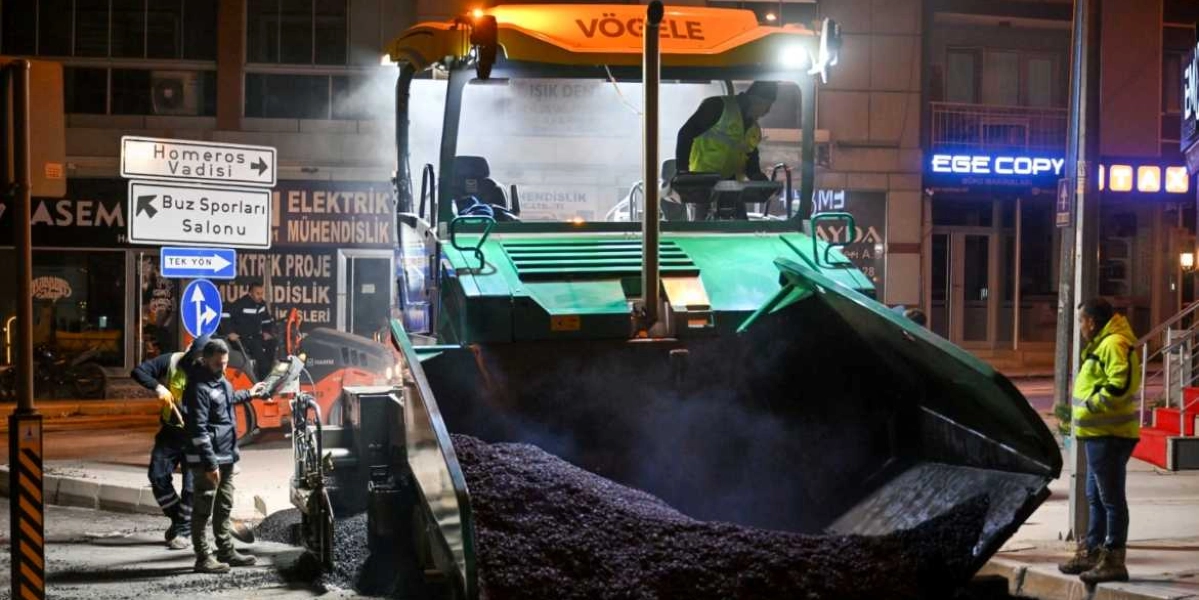 Gece vakti şehir merkezinde çalışan yeşil renkli Vögele marka asfalt serim makinesi ve çevresinde reflektörlü iş kıyafetleri giymiş görevliler. Arka planda dükkan tabelaları, 