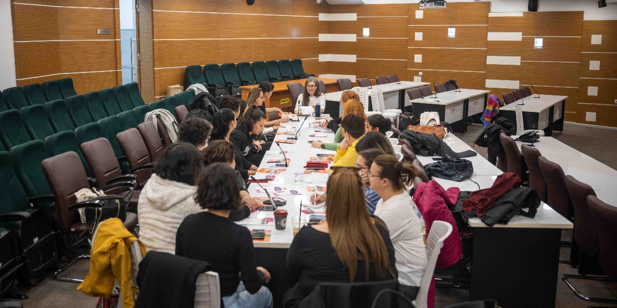 Modern bir toplantı salonunda, uzun bir masa etrafında toplanmış kadınlardan oluşan bir grup, kadınların insan hakları konulu bir çalışma veya toplantı gerçekleştiriyor. Katılımcılar masanın her iki tarafına dizilmiş, önlerinde not defterleri, kalemler ve çeşitli dokümanlar bulunuyor. Salonun duvarları ahşap panellerle kaplı ve arka planda çok sayıda boş koltuk yer alıyor. Masanın en başında oturan bir kişi, elindeki belgeleri incelerken diğer katılımcılar dikkatle dinliyor veya not alıyor.