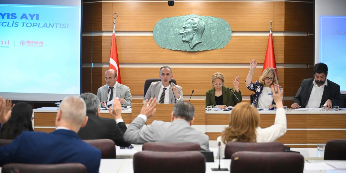 Bornova Belediyesi meclis toplantısında, başkanlık divanında oturan yetkililer ve salondaki meclis üyeleri bir oylama sırasında ellerini kaldırarak oy kullanıyor. Arka planda ahşap paneller üzerinde Mustafa Kemal Atatürk'ün kabartma portresi ve iki yanında Türk bayrakları yer alıyor. Soldaki projeksiyon ekranında 