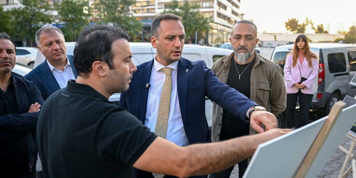 Açık havada, Bornova Belediye Başkanı Mustafa İduğ'un da aralarında bulunduğu bir grup, bir proje panosunu inceliyor. Lacivert ceketli ve desenli kravatlı Mustafa İduğ, eliyle panodaki bir detayı işaret ederek yanındaki kişilere bilgi veriyor. Grubun geri kalanında siyah tişörtlü, ceketli ve günlük kıyafetli kişiler yer alıyor; arka planda ise beyaz bir minibüs ve şehir dokusu görülüyor.