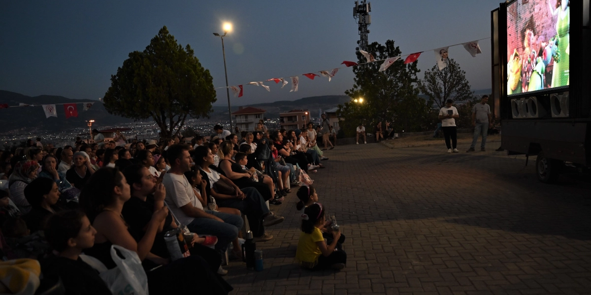 Alacakaranlıkta açık havada kurulan dev bir ekranda çizgi film izleyen kalabalık bir grup insan. Parke taşlı bir alanda oturan izleyicilerin üzerinde Türk bayrakları ve Atatürk görselleriyle süslü flamalar asılı. Sağ tarafta büyük bir ekranın monte edildiği bir kamyonet ve yanında duran görevliler bulunurken, arka planda şehir ışıkları ve ağaçlar yer alıyor.