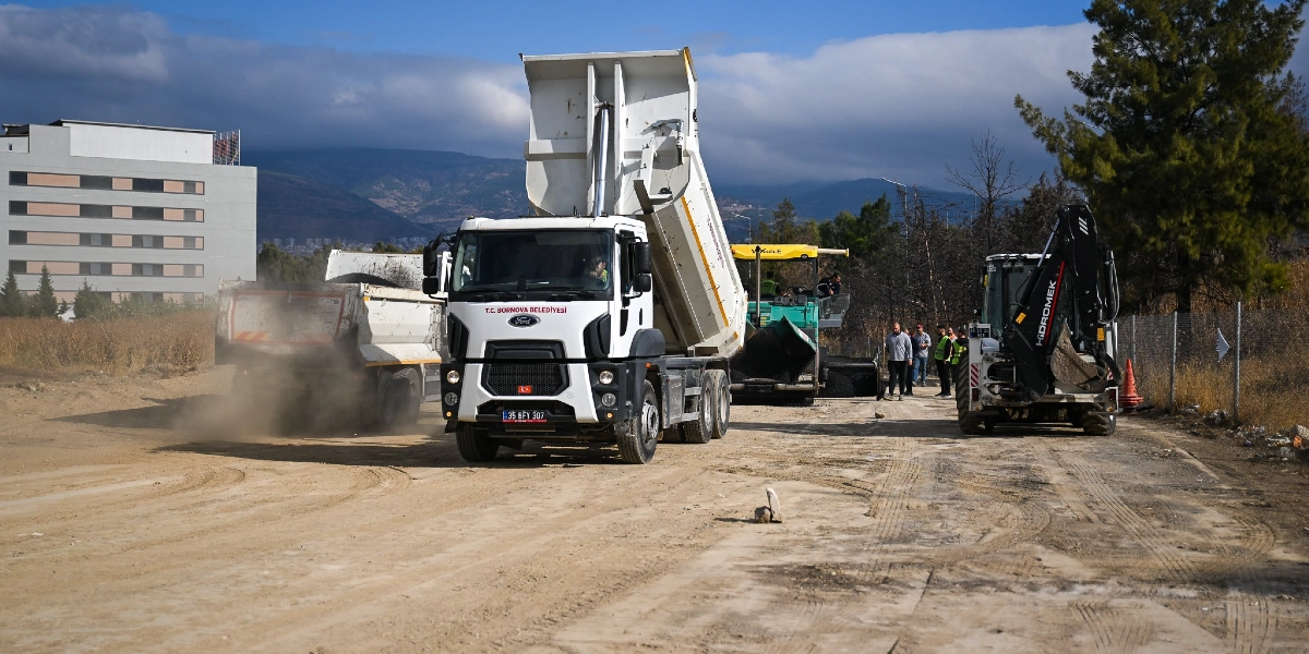 Bornova Belediyesi'ne ait damperli bir kamyonun toprak bir yolda asfalt döküm çalışması yaptığı, yanında bir iş makinesi ve görevlilerin bulunduğu yol yapım sahası.