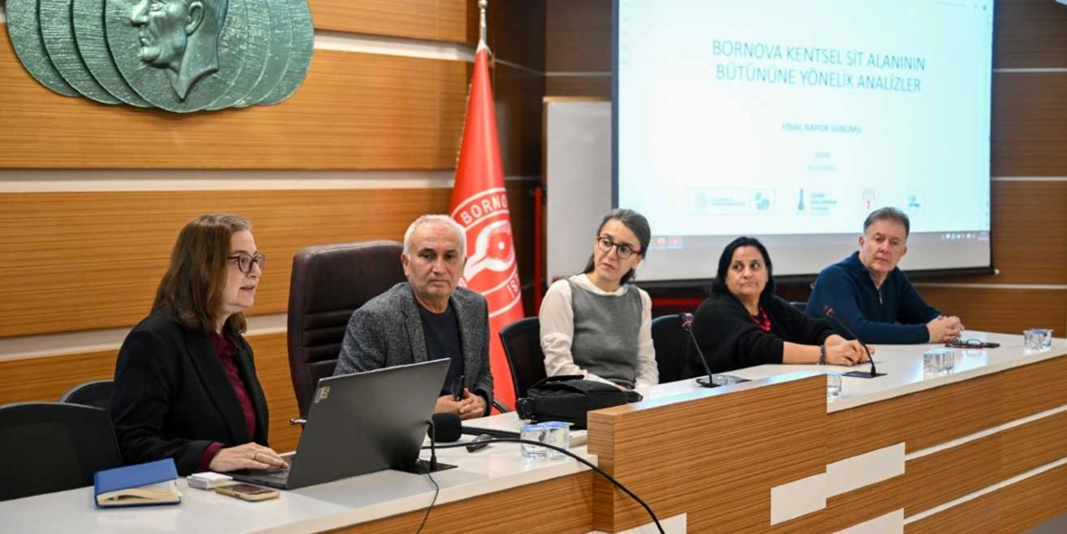 Toplantı masasında yan yana oturan beş kişilik bir grup, Bornova Kentsel Sit Alanı'na yönelik analizlerin sunulduğu bir toplantıda yer alıyor. En solda, önünde dizüstü bilgisayar bulunan bir kadın konuşma yaparken, yanında dört kişi onu dinliyor. Arka planda, üzerinde 