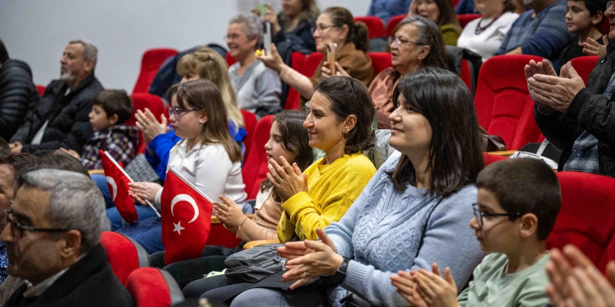 Kırmızı koltuklu bir konser salonunda oturan ve alkışlayan izleyiciler. Ön planda sarı kazaklı bir kadın, yanında açık mavi kazaklı bir kadın ve gözlüklü bir çocuk yer alıyor. İzleyicilerin bir kısmı ellerinde küçük Türk bayrakları tutuyor. Arka sıralarda ise farklı yaş gruplarından insanlar sahneye doğru bakarak alkışlamaya devam ediyor.