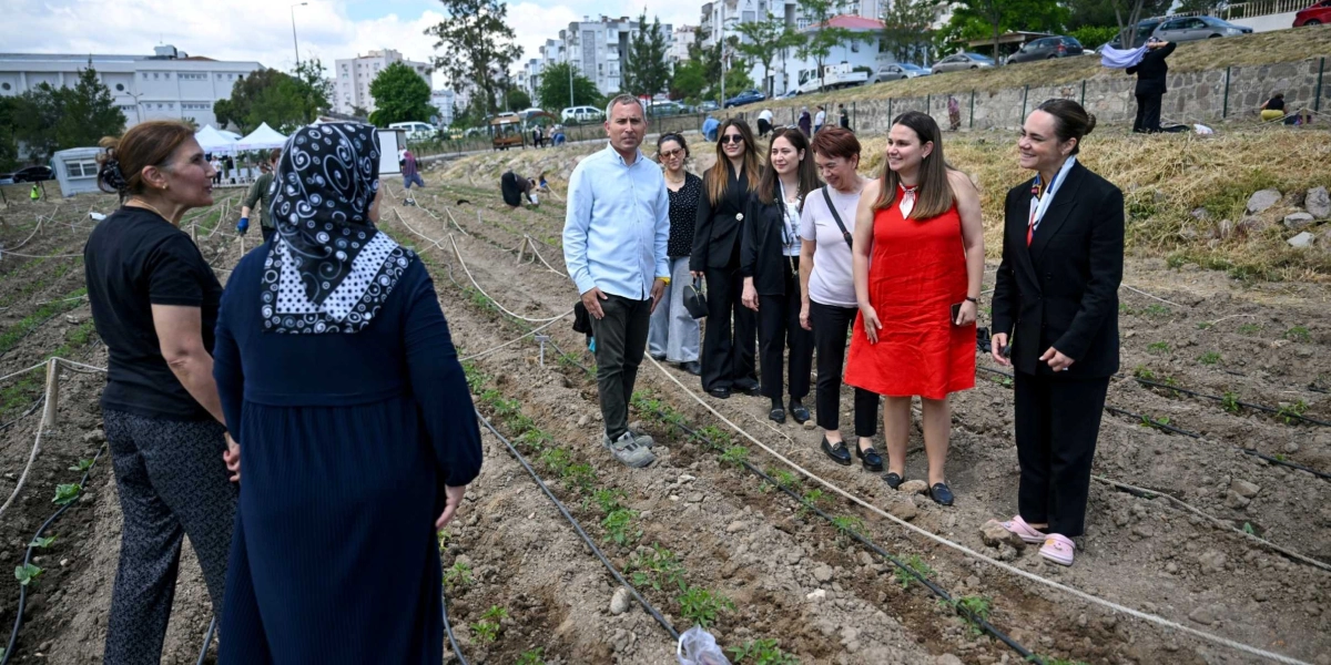 Açık havada, yeni ekilmiş fidelerin bulunduğu bir tarım alanında bir araya gelen bir grup kadın ve bir erkek, toprak üzerinde yürürken sohbet ediyor. Önde, koyu renkli kıyafetler giymiş iki kadın birbirine dönük dururken, arkalarında açık mavi gömlekli bir erkek ve yanındaki beş kadın, ekili alan boyunca yan yana dizilmiş şekilde ilerliyor. Arka planda ise binalar, ağaçlar ve eğimli bir arazi görülüyor.