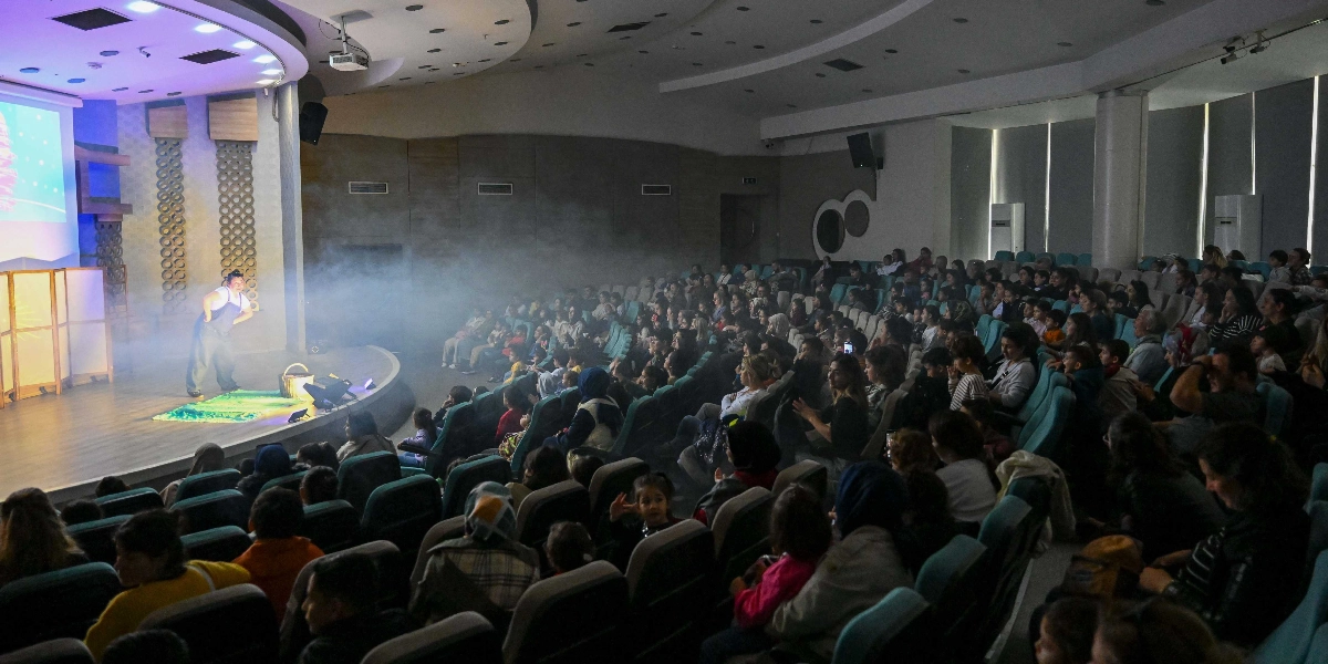 Modern bir tiyatro salonunda sahne üzerinde performans sergileyen bir oyuncu ve onu izleyen kalabalık bir çocuk ve ebeveyn grubu. Sahne, sis efektleriyle aydınlatılmış durumda ve oyuncu, izleyicilere yönelik bir gösteri sunuyor. Salonun koltukları dolu ve izleyiciler sahneye odaklanmış şekilde oturuyor.
