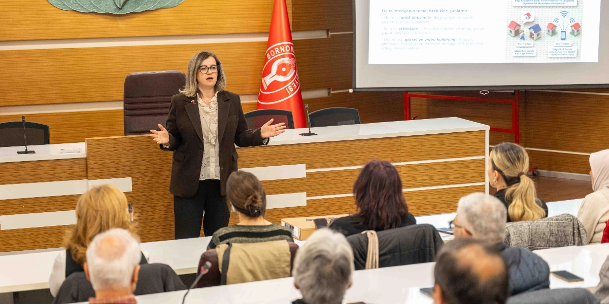 Bornova Belediyesi'ne ait bir toplantı salonunda, gözlüklü ve koyu renk ceketli bir kadın eğitmen, katılımcılara dijital dönüşüm hakkında sunum yapıyor. Eğitmen, kürsü önünde ayakta durarak elleriyle konuşmasını destekliyor. Arkasındaki projeksiyon perdesinde dijital medyanın temel özellikleri ve iletişim yöntemlerini anlatan bir sunum yansıtılıyor. Salonun ön tarafında Bornova Belediyesi'nin kırmızı renkli bayrağı yer alırken, katılımcılar sıralarda oturarak eğitimi takip ediyor.