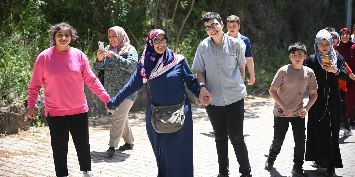 Açık havada, parke taşlı bir yolda el ele tutuşarak yürüyen ve gülümseyen bir grup insan. En önde pembe sweatshirt giyen genç bir kadın, yanında lacivert ferace ve çiçekli eşarp takan orta yaşlı bir kadın, onun yanında ise gri gömlekli genç bir erkek yer alıyor. Grubun arkasında onları cep telefonuyla görüntüleyen başörtülü bir kadın ve yanlarında yürüyen başka gençler bulunuyor.