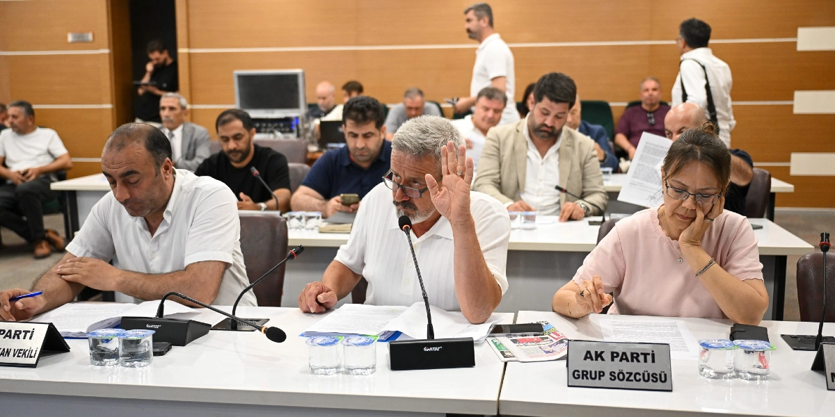 Toplantı masasında oturan bir grup insan, önlerindeki belgeleri inceliyor. Ön planda, beyaz gömlekli, gözlüklü bir katılımcı elini kaldırarak söz istiyor. Yanında beyaz gömlekli bir erkek ve pembe bluzlu bir kadın oturuyor. Masaların üzerinde mikrofonlar, su bardakları ve üzerinde 