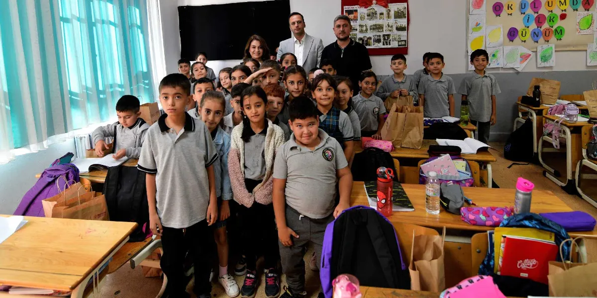 Sınıf ortamında bir araya gelmiş ilkokul öğrencileri ve arkalarında duran yetişkinler, masaların üzerinde duran beslenme çantaları ve okul malzemeleriyle birlikte objektife gülümsüyor.