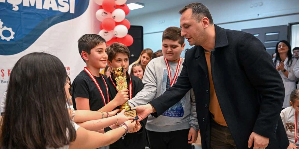 Siyah ceketli bir yetişkin, bir bilgi yarışmasında dereceye giren öğrencilere altın renkli kupalarını takdim ediyor. Öğrenciler boyunlarında madalyalarıyla gülümseyerek kupayı tutarken, arka planda kırmızı ve beyaz balonlarla süslenmiş bir etkinlik alanı ve katılımcılar yer alıyor.