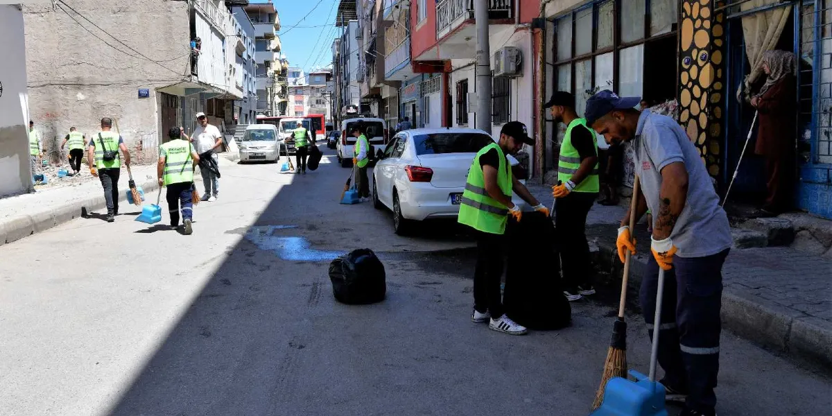 Bornova sokaklarında temizlik yapan, üzerlerinde neon yeşil yelekler bulunan belediye personeli, ellerinde süpürge ve faraşlarla cadde üzerindeki çöpleri topluyor. Bazı çalışanlar süpürdükleri çöpleri siyah çöp poşetlerine doldururken, diğerleri sokağın farklı noktalarında temizlik faaliyetine devam ediyor. Arka planda park halindeki araçlar ve binalar görülüyor.
