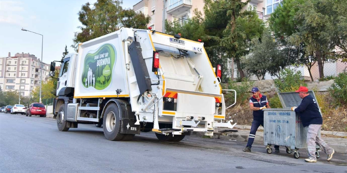 Bornova Belediyesi’ne ait beyaz bir çöp kamyonu, yol kenarında duran metal bir çöp konteynerini boşaltmaya hazırlanan iki belediye işçisiyle birlikte görülüyor. Kamyonun yan tarafında 
