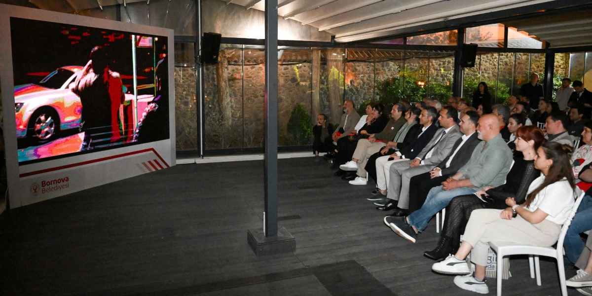 Modern bir mekanda, büyük bir ekrana yansıtılan kısa filmi izleyen kalabalık bir izleyici grubu. Ekranın sol alt köşesinde Bornova Belediyesi logosu yer alıyor. İzleyiciler, cam duvarlarla çevrili, taş duvarlı bir bahçe manzarasına sahip kapalı bir alanda sandalyelerde oturarak ekranı dikkatle takip ediyor.