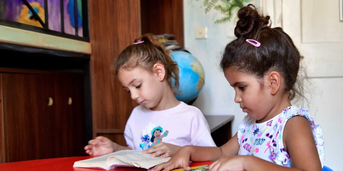 Kütüphane ortamında kırmızı bir masada yan yana oturup kitap okuyan iki küçük kız çocuğu. Soldaki çocuk açık renkli, üzerinde çizgi film karakteri baskısı olan bir tişört giyiyor ve saçları at kuyruğu şeklinde toplanmış. Sağdaki çocuk ise üzerinde renkli desenler bulunan kolsuz bir bluz giyiyor ve saçları tepeden topuz yapılmış. Arka planda ahşap bir kitaplık ve üzerinde dünya küresi bulunuyor.