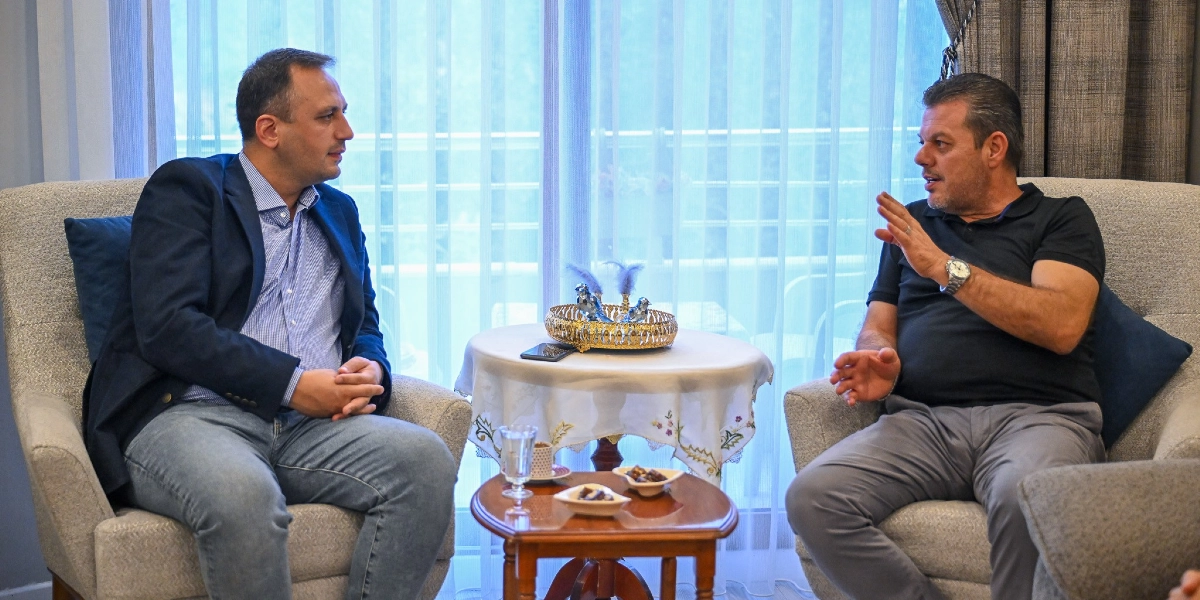 Lacivert ceketli ve açık mavi gömlekli bir erkek ile siyah polo yaka tişörtlü bir erkek, karşılıklı koltuklarda oturarak sohbet ediyor. Aralarındaki küçük sehpanın üzerinde bir bardak su ve atıştırmalıklar bulunuyor.