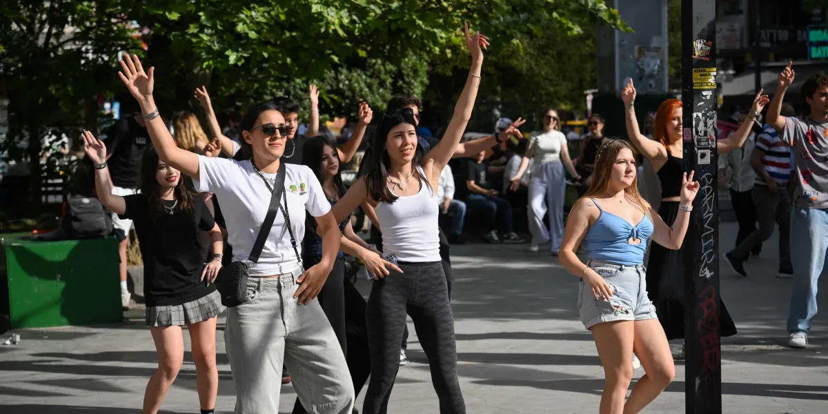Bornova sokaklarında güneşli bir günde, kollarını havaya kaldırarak neşeyle dans eden ve eğlenen bir grup genç kadın. Ön planda beyaz tişörtlü, güneş gözlüklü bir kadın, yanında beyaz atletli bir başka kadın ve mavi büstiyerli, kot şortlu bir genç kadın yer alıyor. Arka planda ise ağaçların gölgesinde dansa eşlik eden diğer gençler ve yürüyen insanlar görünüyor.