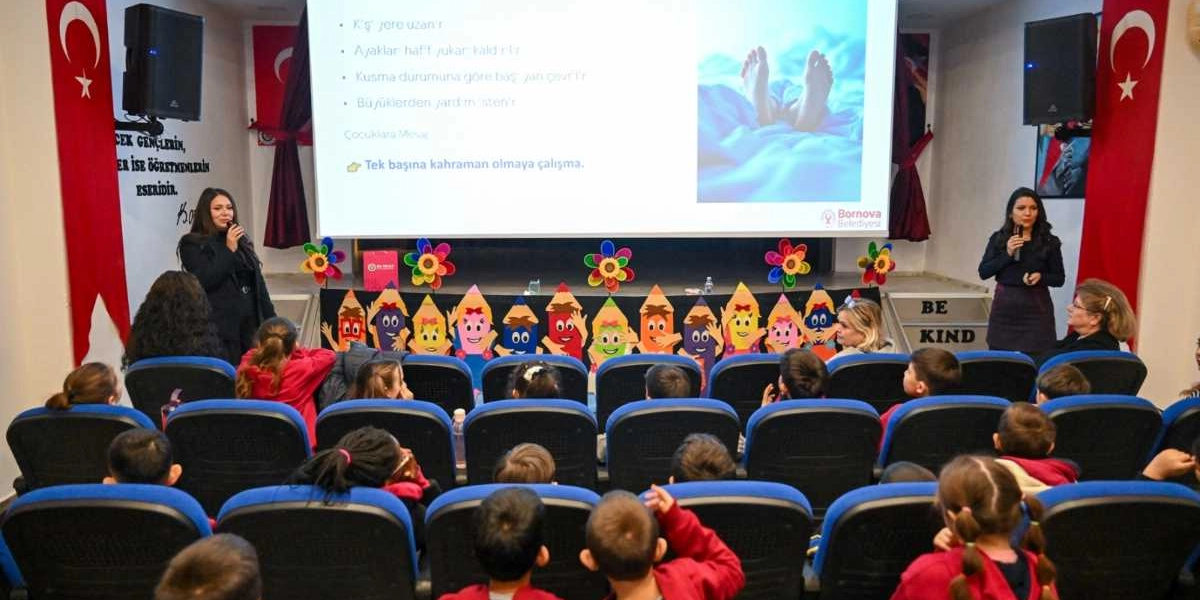 Bir okulun konferans salonunda, ilkokul ve ortaokul öğrencilerine yönelik düzenlenen sağlık eğitiminde, iki kadın eğitmen mikrofonla sunum yapıyor. Salonun önündeki büyük ekranda temel ilk yardım bilgileri ve 