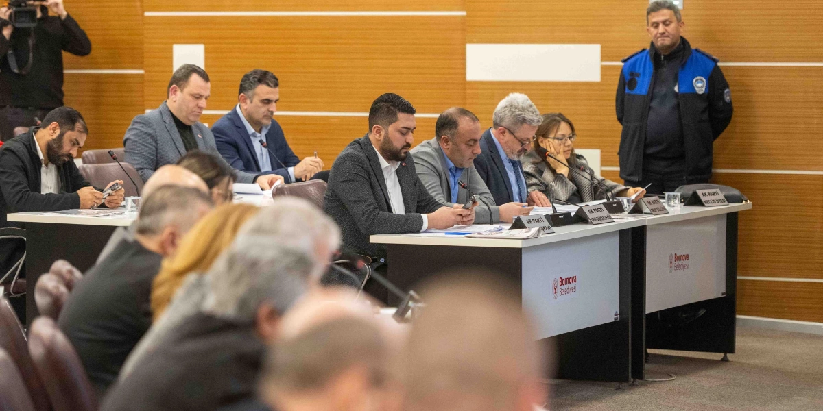 Bornova Belediyesi meclis salonunda, masalarda oturan ve önlerinde Bornova Belediyesi logolu isimlikler bulunan bir grup katılımcı toplantı yapıyor. Katılımcıların çoğu takım elbiseli olup önlerindeki belgelere bakıyor veya telefonlarıyla ilgileniyor. Arka planda ayakta duran bir güvenlik görevlisi ve sol tarafta elinde kamera tutan bir kişi yer alıyor.