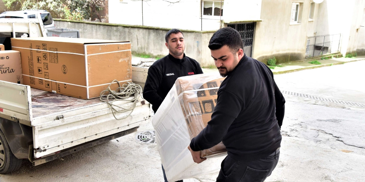 Üzerinde Regal ve Vestel logolu siyah üniformalar bulunan iki genç, bir kamyonetin kasasından indirdikleri paketlenmiş beyaz eşyayı taşıyorlar. Kamyonetin kasasında başka büyük karton kutular da bulunuyor.