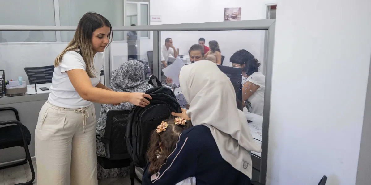 Beyaz tişörtlü ve bej pantolonlu bir kadın, ofis ortamında yanında duran başörtülü bir anne ve küçük kızına siyah bir okul çantası uzatıyor. Arka planda cam bölmelerin ardında çalışan diğer personel ve işlem bekleyen insanlar görünüyor.