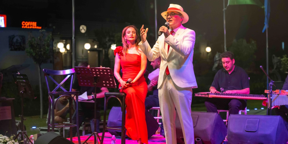 Gece düzenlenen bir etkinlikte, beyaz takım elbiseli ve şapkalı bir erkek şarkıcı mikrofonla şarkı söylerken, yanında kırmızı tek omuzlu abiye elbise giymiş bir kadın sanatçı oturuyor. Sahnenin arka planında ise kanun çalan bir müzisyen ve diğer orkestra üyeleri yer alıyor.
