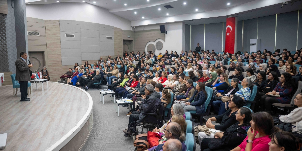 Modern bir konferans salonunda, kürsüde elinde mikrofonla konuşma yapan bir erkek ve onu dikkatle dinleyen kalabalık bir dinleyici grubu. Salonun ön kısmında sahne üzerinde konuşmacı yer alırken, izleyiciler kademeli olarak yerleştirilmiş turkuaz renkli koltuklarda oturuyor. Arka planda duvarda asılı bir Türk bayrağı bulunuyor ve katılımcıların çoğunluğunu kadınların oluşturduğu geniş bir kitle etkinliği takip ediyor.