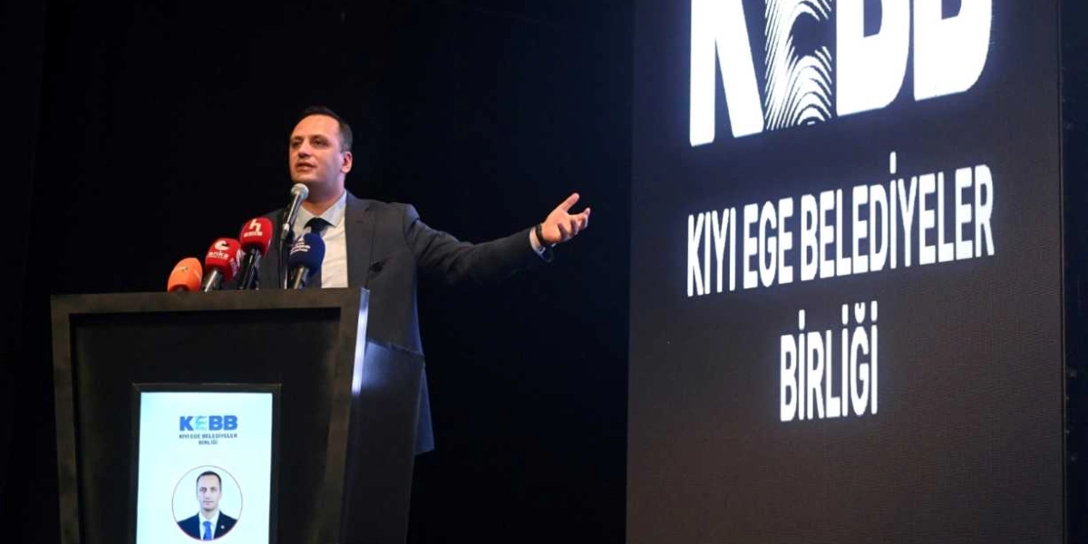 Koyu renkli bir sahnede, kürsüde konuşma yapan bir erkek, sol eliyle izleyicilere hitap ediyor. Kürsünün ön yüzünde Kıyı Ege Belediyeler Birliği logosu ve konuşmacının fotoğrafı yer alıyor. Sağ taraftaki büyük dijital ekranda ise parmak izi görseliyle birleştirilmiş 