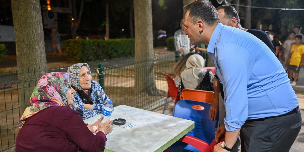 Açık havada, ağaçlıklı bir park ortamında, mavi gömlekli bir erkek, masada oturan iki yaşlı kadınla sohbet ediyor. Kadınlardan biri elinde bir su şişesi tutarken, diğeri ona dikkatle bakıyor. Arka planda ise ayakta duran diğer insanlar ve parkın çitleri görülüyor.