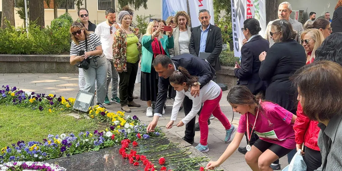 Bornova'da düzenlenen Anneler Günü etkinliğinde, bir grup çocuk ve yetişkin Atatürk Anıtı'nın önüne kırmızı karanfiller bırakıyor. Ön planda pembe tişörtlü bir kız çocuğu ve yanında beyaz tişörtlü, pembe pantolonlu başka bir kız çocuğu ile ona eşlik eden bir erkek, anıta çiçeklerini yerleştiriyor. Çevredeki kalabalık grup, bu anı izliyor ve bazıları cep telefonlarıyla fotoğraf çekiyor. Arka planda ağaçlar ve Bornova Belediyesi'ne ait flamalar görülüyor.