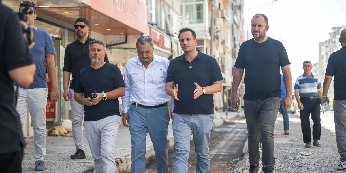 Mustafa Kemal Caddesi'nde devam eden yol yenileme çalışmaları sırasında, aralarında yerel yöneticilerin de bulunduğu bir grup erkek, cadde üzerinde yürüyerek incelemelerde bulunuyor. Öndeki grupta yer alan beyaz gömlekli ve açık renk pantolonlu bir kişi ile yanındaki lacivert tişörtlü kişi konuşarak ilerliyor. Arka planda ise dükkanların bulunduğu cadde ve kaldırımda dinlenen bir köpek görülüyor.