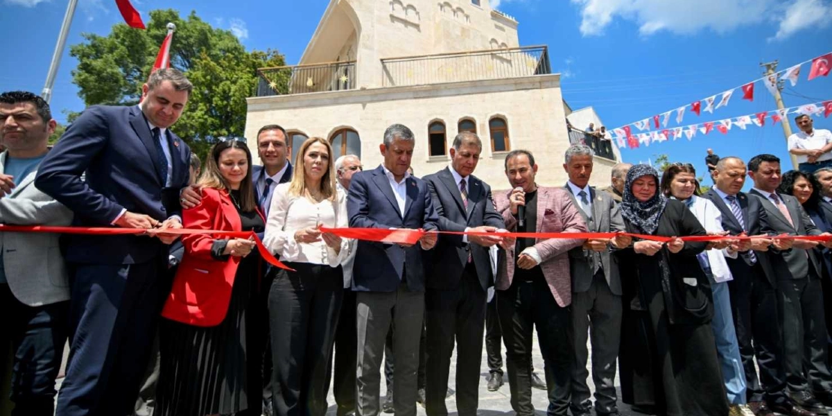 CHP Genel Başkanı Özgür Özel ve beraberindeki heyet, Bornova'da yeni bir tesisin açılış kurdelesini kesiyor. Aralarında yerel yöneticilerin de bulunduğu grup, taş mimariye sahip binanın önünde yan yana dizilmiş, ellerindeki makaslarla kırmızı kurdeleyi tutuyor. Açık havada, güneşli bir günde gerçekleşen törende katılımcılar resmi kıyafetler giymiş durumda ve arka planda Türk bayrakları ile süslemeler yer alıyor.