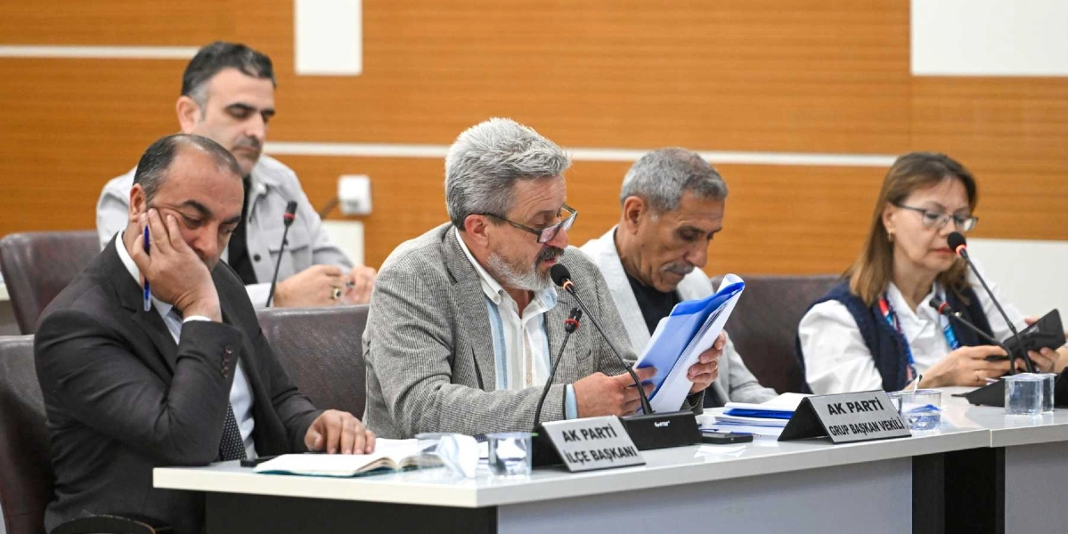 Bornova Belediyesi meclis toplantısında, masada oturan dört kişilik bir grup yer alıyor. Ortada oturan gözlüklü ve gri ceketli kişi elindeki dosyayı incelerken, yanında 