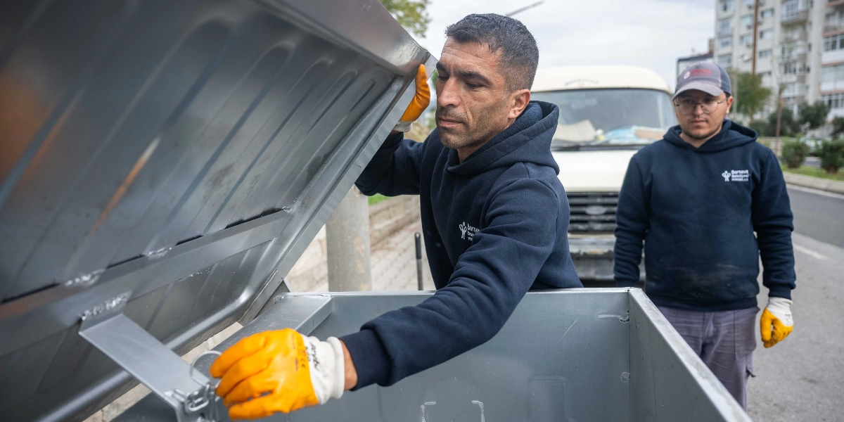 Bornova Belediyesi logolu lacivert sweatshirt giyen iki belediye çalışanı, sokak üzerinde yeni metal çöp konteynerlerini yerleştiriyor. Öndeki çalışan konteynerin kapağını açarken, arkadaki çalışan onu izliyor. Her iki görevli de sarı iş eldivenleri takıyor.