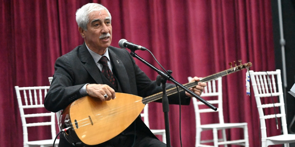 Koyu renk takım elbiseli, beyaz saçlı ve bıyıklı bir sanatçı, elinde sazıyla mikrofon başında şarkı söylüyor. Arka planda kırmızı perde ve beyaz sandalyeler bulunuyor.
