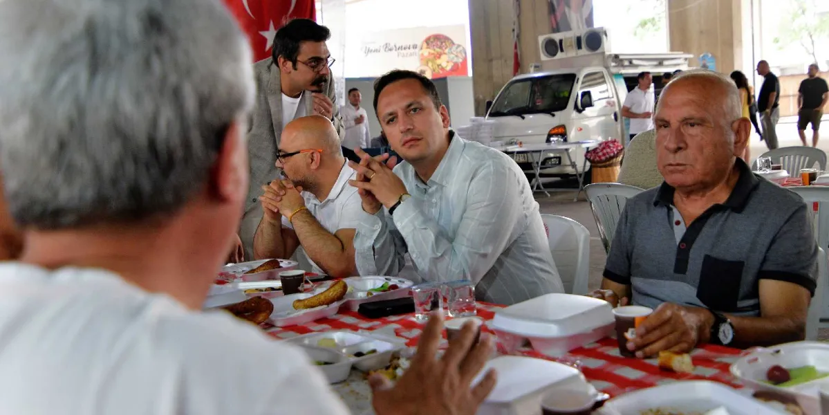 Kapalı bir alanda, kırmızı beyaz kareli örtü serili uzun bir masada oturan bir grup erkek, önlerindeki yemek tabakları ve içeceklerle sohbet ediyor. Masanın merkezinde açık renkli gömlek giymiş bir kişi ellerini kavuşturmuş dikkatle dinlerken, yanında gözlüklü ve beyaz gömlekli bir kişi ile sağ tarafta gri polo yaka tişörtlü yaşlı bir bey yer alıyor. Arka planda ise üzerinde 