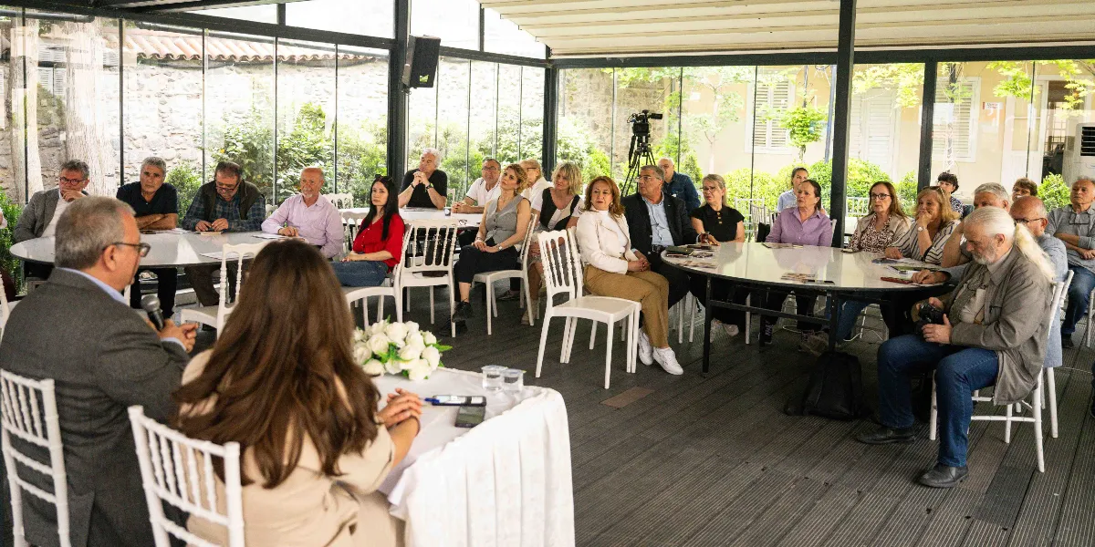 Cam duvarlarla çevrili, aydınlık bir mekanda düzenlenen toplantıda, masaların etrafında oturan yaklaşık yirmi kişilik bir grup yer alıyor. Ön planda, sırtı kameraya dönük uzun saçlı bir kadın ve yanında konuşma yapan gri saçlı bir adam bulunuyor. Katılımcıların çoğu masalarda oturarak dinleyici konumunda yer alırken, sağ tarafta elinde fotoğraf makinesi tutan beyaz saçlı ve sakallı bir adam dikkat çekiyor. Mekanın arka planında ağaçlar ve dış mekan görünürken, ortamda resmi bir toplantı veya panel havası hakim.