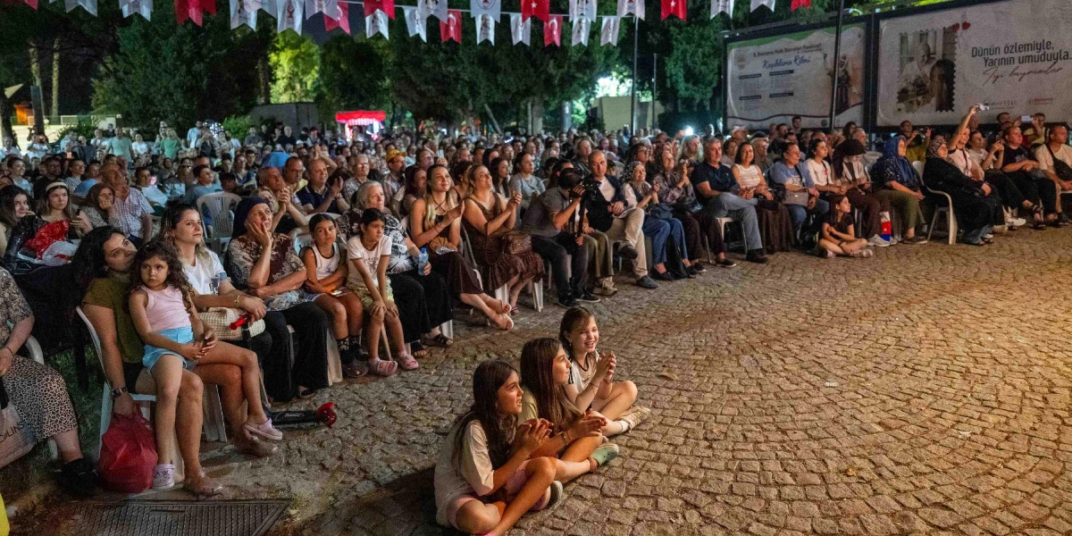 Açık havada, taş döşeli bir alanda düzenlenen etkinliği izleyen kalabalık bir izleyici grubu. Ön planda yere oturmuş alkışlayan iki kız çocuğu, hemen arkalarında ise sandalyelerde oturan çocuklar, gençler ve yetişkinler yer alıyor. Etkinlik alanı, üzerinde Türk bayrakları ve çeşitli flamaların asılı olduğu iplerle süslenmiş. Arka planda ise üzerinde 