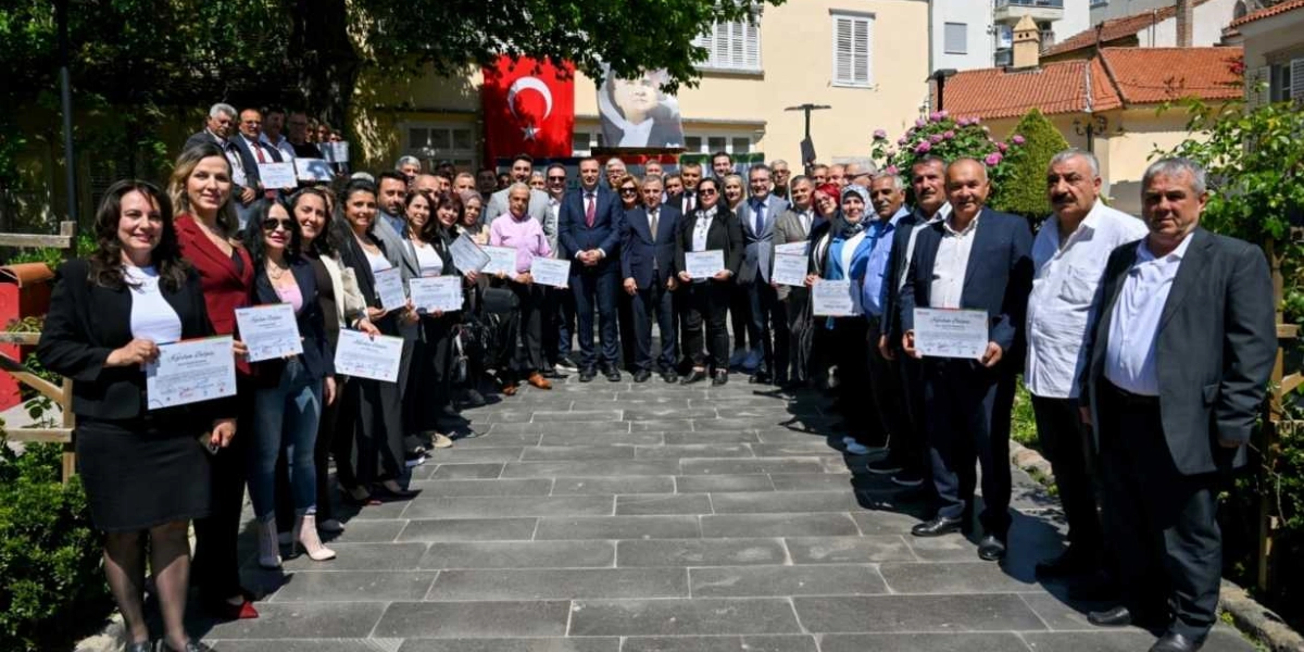 Açık havada, taş döşeli bir yolda yan yana dizilmiş, ellerinde katılım belgeleri tutan kalabalık bir grup insan. Arka planda tarihi bir bina, binanın üzerinde asılı Türk bayrağı ve Mustafa Kemal Atatürk'ün portresi yer alıyor. Resmi kıyafetli kadın ve erkeklerden oluşan grup, objektife gülümseyerek poz veriyor.
