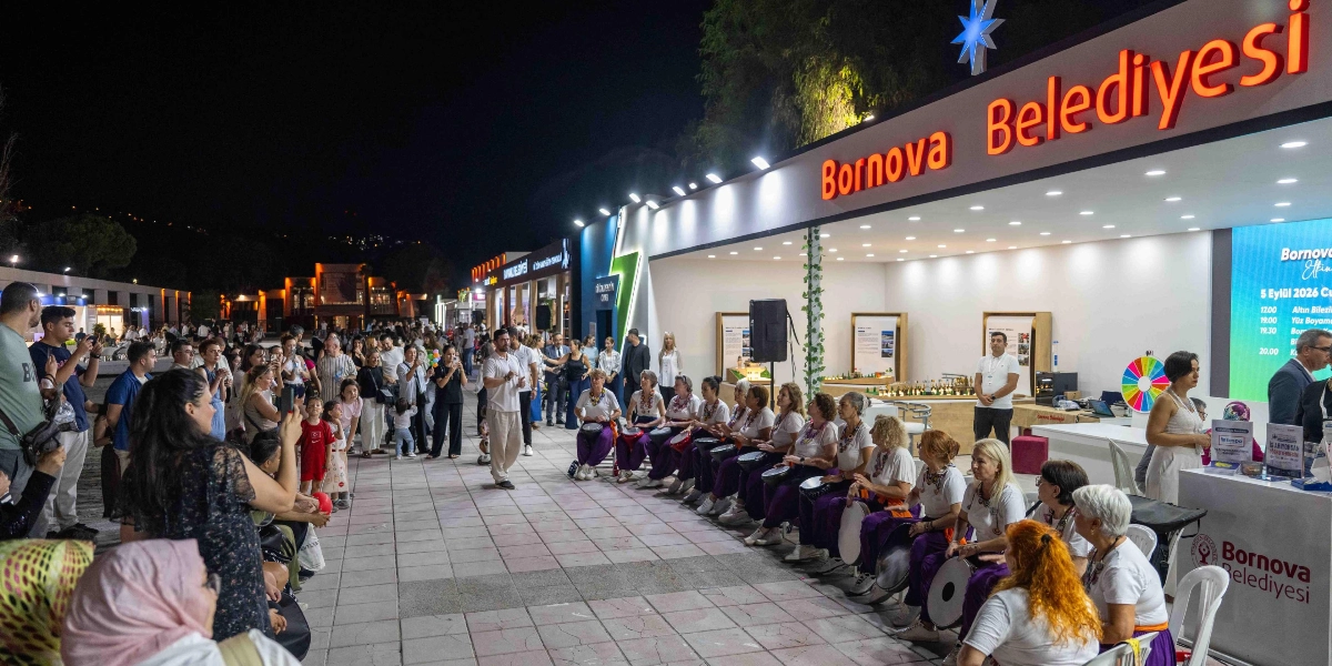 İzmir Enternasyonal Fuarı alanında, gece vakti Bornova Belediyesi standının önünde düzenlenen etkinlikte, beyaz tişörtlü ve mor pantolonlu kadınlardan oluşan bir perküsyon grubu ellerindeki davullarla performans sergiliyor. Grubun önünde bir eğitmen yönlendirme yaparken, çevrede toplanan kalabalık grubu izliyor ve bazıları cep telefonlarıyla kayıt alıyor. Standın üzerinde ışıklı 
