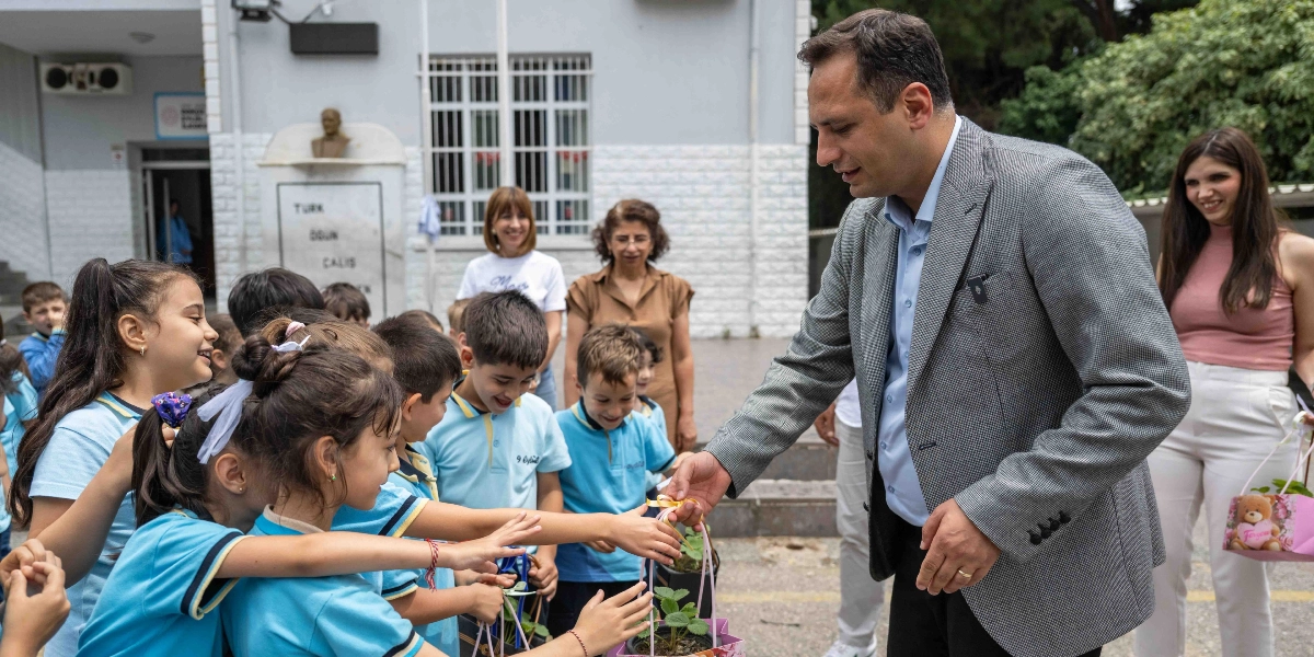 Okul bahçesinde, üzerinde açık mavi okul formaları olan ilkokul öğrencileri, ellerinde küçük saksılarda fide taşıyan bir yetişkinle etkileşim halindeler. Ceketli bir erkek, çocuklara saksıları uzatırken, arkada öğretmenler ve okul binası yer alıyor. Çocuklar heyecanla fideleri inceliyor ve toprakla buluşturmaya hazırlanıyorlar.
