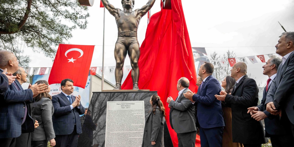 Bornova Pınarbaşı'nda düzenlenen törenle açılan Naim Süleymanoğlu heykeli, halterciyi başının üzerinde ağırlık kaldırırken tasvir ediyor. Bronz renkli heykelin kaidesinde, üzerinde Naim Süleymanoğlu'nun biyografisinin yer aldığı mermer bir plaket bulunuyor. Heykelin açılışını yapan protokol üyeleri ve katılımcılar, heykelin iki yanında durarak alkışlıyor; arka planda Türk bayrakları ve süslemeler yer alıyor.