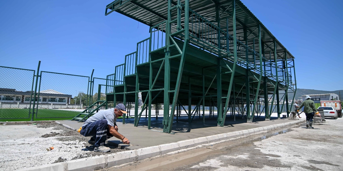 Açık havada, yeni inşa edilen yeşil metal tribün yapısının önünde bir işçi, elindeki mala ile taze dökülmüş beton zemini düzeltiyor. Arka planda tel örgülerle çevrili bir spor sahası ve başka işçiler görülüyor.